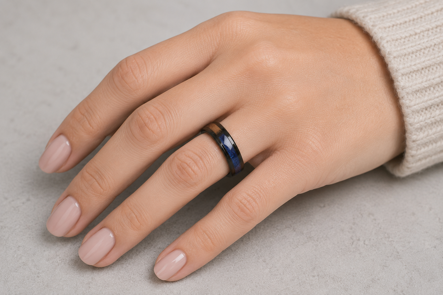 A close-up of a hand wearing a dark metallic ring with a polished gold accent stripe, resting on a light gray textured surface.